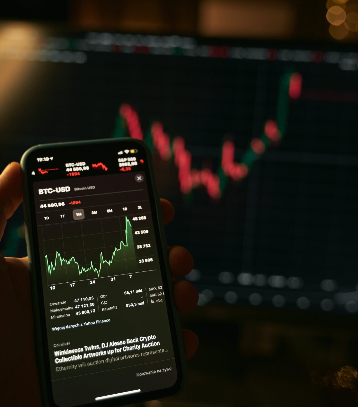 Hand holding a modern smartphone with the stocks market app and big screen blurred on the background
